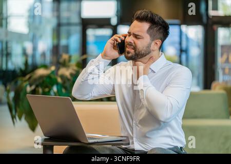 Homme d'affaires malade parlant sur smartphone ayant mal à la gorge, toux et travaille avec un ordinateur portable. Homme fatigué stressé souffrant de rhume ou de grippe travaillant dans le hall du bureau. Triste travailleur a eu virus, maladie. Banque D'Images