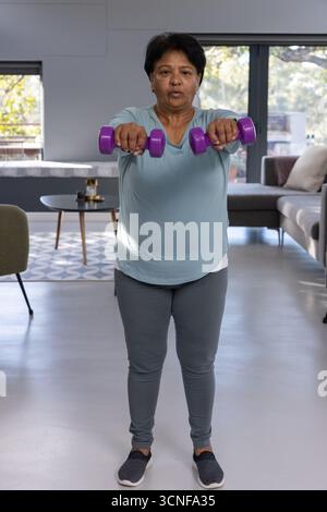 Femme indienne senior soulevant des haltères violets dans le salon portant un haut et des leggings gris Banque D'Images