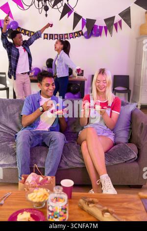 Divers amis assis sur le canapé gris manger du gâteau et danser dans le salon, bannière d'anniversaire Banque D'Images