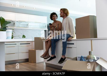 Couple diversifié assis sur le bord de l'île dans la cuisine déballant les boîtes de déménagement, tenant smartphone Banque D'Images
