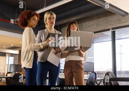 Diverses collègues féminines debout dans le bureau examinant la tablette portable et le document imprimé Banque D'Images