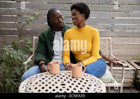 Couple afro-américain assis et bavardant sur la terrasse en bois autour d'une table en métal tenant des tasses à café Banque D'Images