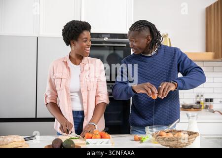 Couple afro-américain tranchant des tomates sur le comptoir en marbre, craquant l'œuf sur le bol dans la cuisine Banque D'Images