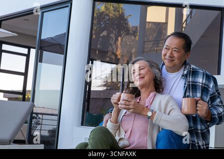 Couple senior diversifié se relaxant sur le balcon patio tenant des tasses près de la fenêtre en verre, espace de copie Banque D'Images