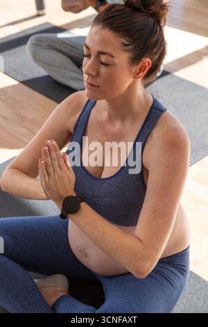 Femme enceinte blanche méditant sur un tapis de yoga gris en studio sur plancher en bois portant une montre-bracelet noire Banque D'Images