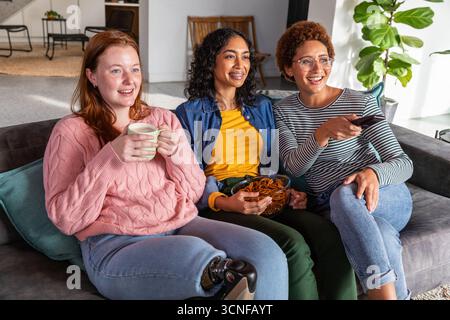 Diverses amies féminines assis sur le canapé dans le salon tenant la tasse, partageant des bretzels avec la télécommande Banque D'Images