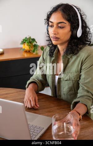 Femme asiatique assise à table dans le bureau à domicile portant des écouteurs eau potable, travaillant sur ordinateur portable Banque D'Images