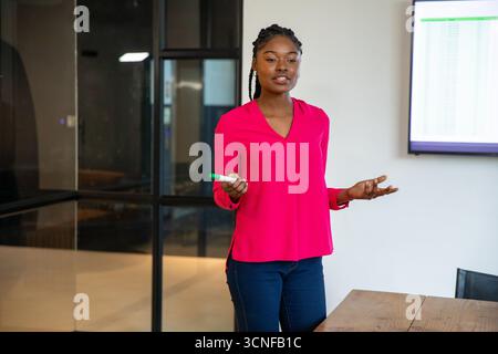 Femme afro-américaine présentant des données de tableur dans une salle de conférence tenant un marqueur à table Banque D'Images