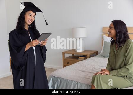 Maman diversifiée et fille adolescente debout et assis dans la chambre portant mortarboard, robe Banque D'Images