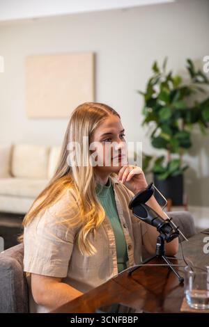 Femme créatrice de contenu enregistrant podcast à table dans le salon avec microphone, verre d'eau Banque D'Images
