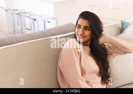 Femme indienne se relaxant sur un canapé beige dans un salon ouvert avec îlot de cuisine et tabourets de bar Banque D'Images