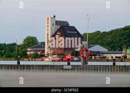Sur une jetée en bois, une bouée de sauvetage orange vif est suspendue à un poteau métallique, le panneau indique bis 38 Fuss signifiant jusqu'à 38 pieds, au loin une bui industrielle Banque D'Images