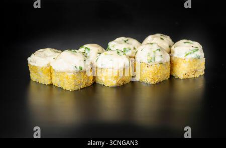 Un gros plan de rouleaux de sushi disposés soigneusement sur une surface noire. Les rouleaux sont recouverts d'une sauce crémeuse et garnis d'herbes vertes. Banque D'Images