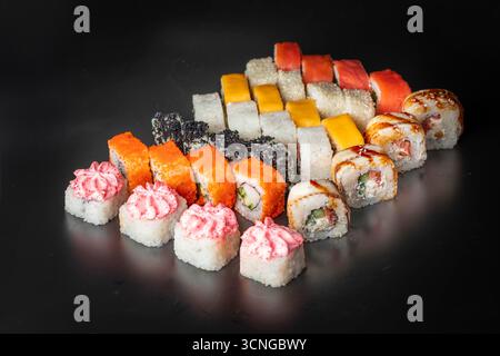 Une variété de rouleaux de sushi disposés sur une surface noire. Comprend des rouleaux colorés avec du poisson, des légumes et des garnitures roses. Idéal pour les menus de restaurant. Banque D'Images