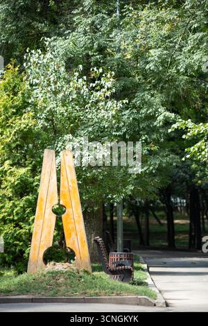 Banc en bois en forme de pince à linge dans un parc. Arbres verts, ombre et atmosphère calme un jour d'été. Banque D'Images