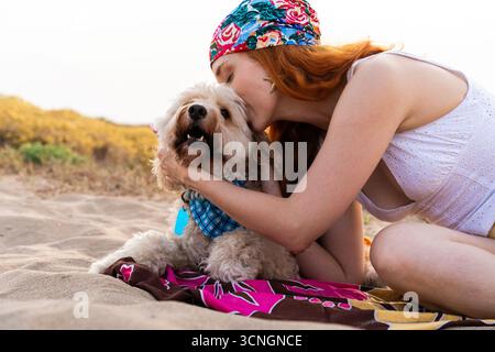 Femme rousse embrassant son chien doodle sur une plage de sable ensoleillée, partageant un moment tendre d'affection et de joie alors qu'ils se détendent ensemble sur une chaude journée d'été Banque D'Images