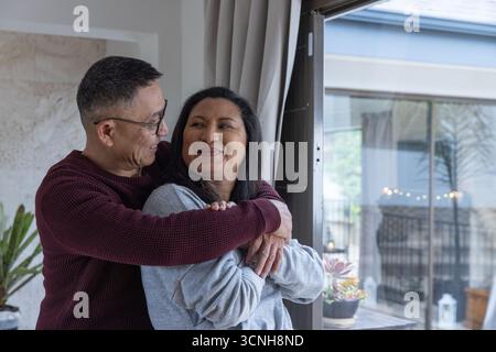 Couple senior embrassant dans le salon avec mur de pierre et plante en pot à côté de la porte coulissante en verre Banque D'Images