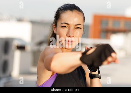 Jeune femme portant des gants pratiquant la boxe ombragée en plein air. Portrait d'une sportive regardant l'appareil photo se réchauffer. Banque D'Images