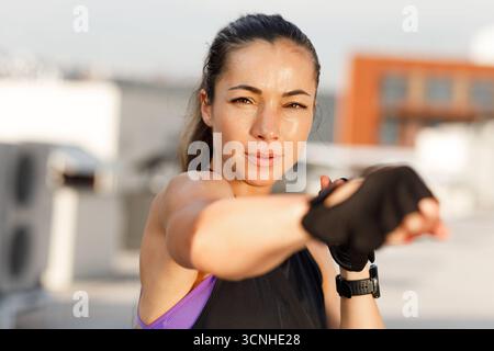 Jeune femme portant des gants pratiquant la boxe ombragée en plein air. Portrait d'une sportive regardant l'appareil photo se réchauffer. Jeune femme portant des gants pract Banque D'Images