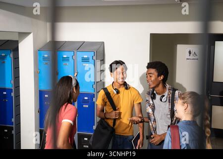 Divers amis discutant près des casiers dans le couloir de l'école tenant une tablette et portant des écouteurs Banque D'Images