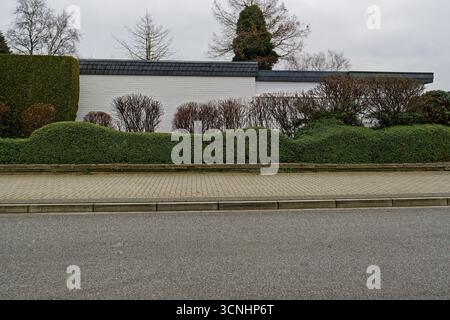 Un trottoir lisse borde une rue calme, une haie vivante taillée de vagues douces coule à côté du trottoir, des bâtiments blancs se tiennent derrière dans une lumière douce, le PA Banque D'Images