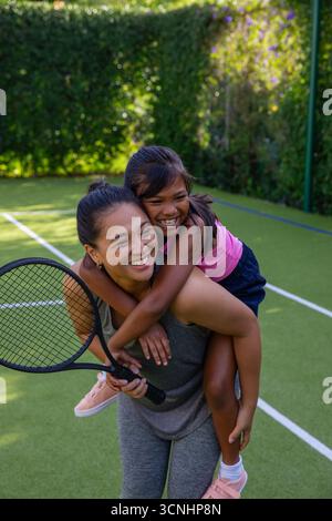 Mère asiatique et fille piggyback sur un court de tennis avec des lignes de limite tenant la raquette dans des chaussures Banque D'Images