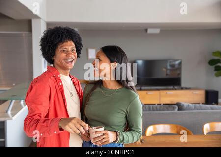 Couple diversifié debout par une table à manger en bois, tenant des tasses en céramique, souriant ensemble à la maison Banque D'Images