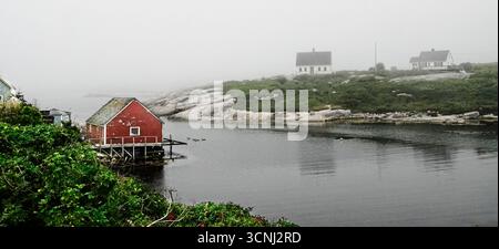 Une scène côtière sereine à Peggy's Cove, en Nouvelle-Écosse, capture un hangar à bateaux rouge niché au bord de l'eau, entouré de brouillard. De vieilles maisons parsèment le paysage Banque D'Images