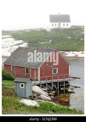 Une scène côtière sereine à Peggy's Cove, en Nouvelle-Écosse, capture un hangar à bateaux rouge niché au bord de l'eau, entouré de brouillard. De vieilles maisons parsèment le paysage Banque D'Images