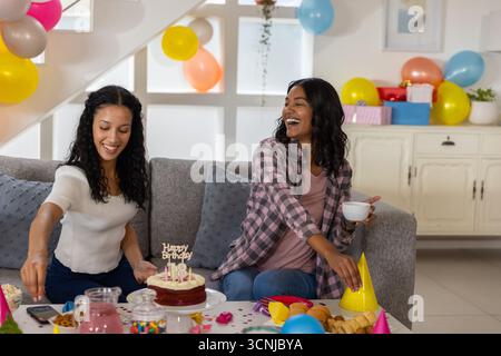 Maman diversifiée et fille adolescente assis sur le canapé gris dans le salon partageant le gâteau 18ème anniversaire Banque D'Images