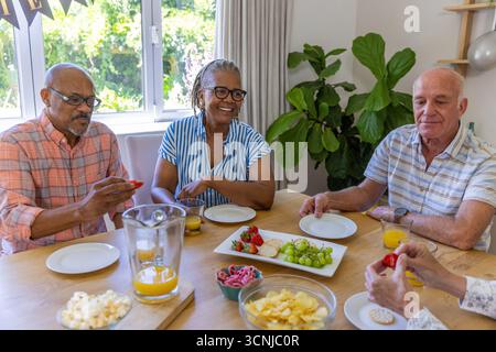 Divers amis seniors assis autour d'une table à manger en bois à la maison, sirotant du jus d'orange, grignotant Banque D'Images
