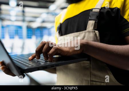 Ingénieur afro-américain ajuste les paramètres industriels pour la production d'énergie solaire, travaillant dans une usine de fabrication de haute technologie. Expert analysant les données techniques sur pc, finetuning. Banque D'Images