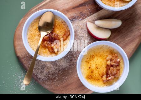 Délicieuse crème brûlée avec une croûte de sucre caramélisée dorée, présentée dans des ramequins élégants sur une surface en bois, complétée par des tranches de pommes fraîches et Banque D'Images