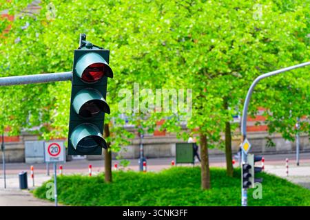 Un triple feu de circulation montre une flèche rouge de virage à gauche, lentilles empilées pour le rouge, l'ambre et le vert. Un panneau de zone vingt se dresse en arrière-plan, arbres et h. Banque D'Images