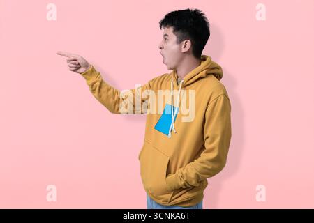 Un jeune homme joyeux portant un sweat à capuche moutarde pointant sur copyspace, placé sur un fond rose doux Banque D'Images