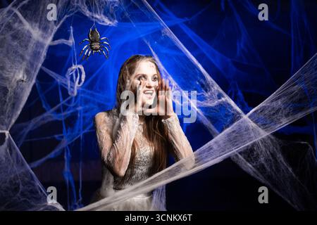 Femme sinistre dans la robe sale blanche avec le maquillage horrible de zombie Halloween criant et faisant des gestes, faisant l'annonce debout sur fond bleu surr Banque D'Images