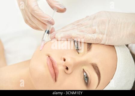 Femme recevant un traitement par injection faciale à mains gantées en milieu médical. Banque D'Images