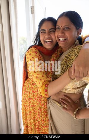 Mère indienne avec fille embrassant et riant par porte coulissante en verre portant saree et kurta Banque D'Images
