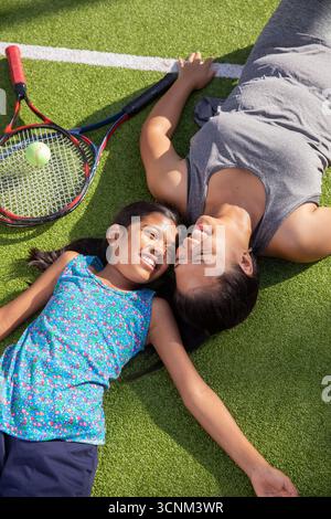 Mère avec fille couchée ensemble sur un court de tennis souriant l'un à l'autre avec des raquettes et balle Banque D'Images