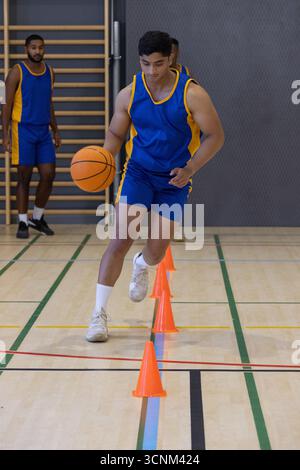 Divers coéquipiers masculins pratiquant l'exercice dribble dans le gymnase avec basket-ball et cônes Banque D'Images