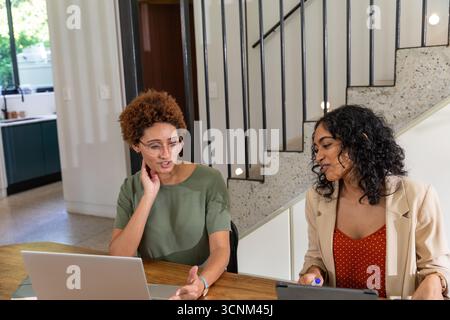Diverses collègues féminines collaborant à la table en bois à la maison, en utilisant un ordinateur portable et une tablette Banque D'Images