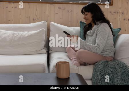 Femme assise sur les jambes croisées sur le canapé dans le salon avec lancer tenant smartphone, espace de copie Banque D'Images