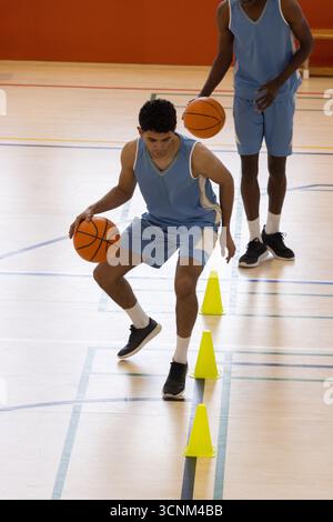 Divers coéquipiers masculins en maillots bleus pratiquant le dribble sur le terrain avec des cornets de basket-ball et des cônes Banque D'Images
