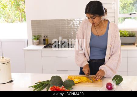 Femme indienne hachant des légumes sur une planche à découper en bois près de la fenêtre dans la cuisine moderne, espace de copie Banque D'Images
