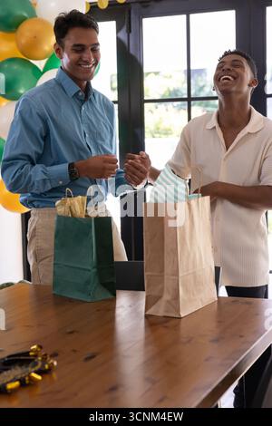 Divers amis masculins debout derrière la table en bois dans la salle à manger près de deux sacs cadeaux, ballons Banque D'Images