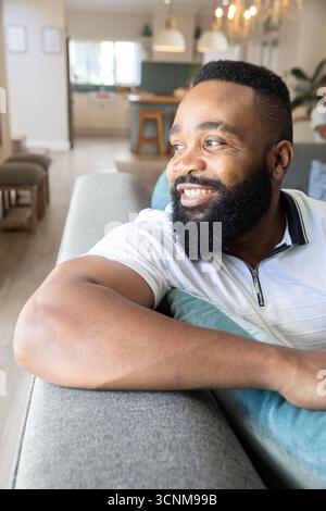 Homme afro-américain appuyé sur le canapé à côté du coussin sarcelle dans le salon décloisonné, espace de copie Banque D'Images