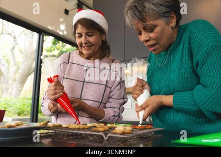 Diverses femmes âgées dans le chapeau de Père Noël passepoil coloré glaçage sur les supports à biscuits en fil au comptoir de cuisine Banque D'Images