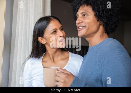 Couple diversifié tenant une tasse en céramique et parlant par fenêtre avec des rideaux à la maison Banque D'Images