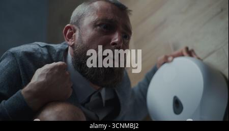 Homme d'affaires avec expression faciale tendue souffrant de diarrhée et d'indigestion, assis sur les toilettes. Homme éprouvant des maux d'estomac, inconfort digestif dans les toilettes du bureau pendant le travail. Gros plan. Banque D'Images