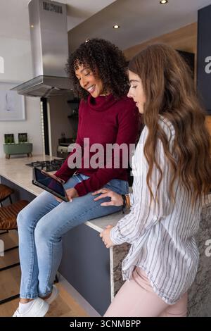 Diverses amies féminines assises sur l'île de marbre avec des tabourets de bar en dessous, examinant la tablette dans la cuisine Banque D'Images